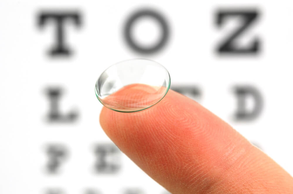 Finger holding up a contact lens in front of an eye exam chart